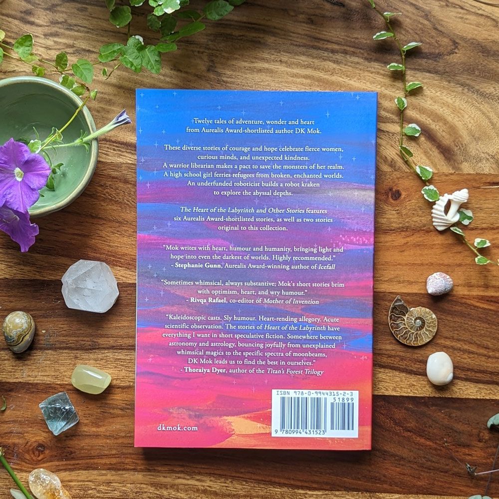 A paperback copy of The Heart of the Labyrinth and Other Stories by DK Mok, resting face-down on a timber desk, surrounded by green leaves, fresh flowers, seashells, and polished stones. The back cover carries a description of the collection: Twelve tales of adventure, wonder and heart from Aurealis Award-shortlisted author DK Mok. These diverse stories of courage and hope celebrate fierce women, curious minds, and unexpected kindness. Beneath the description are three quotes from authors Stephanie Gunn, Rivqa Rafael and Thoraiya Dyer.