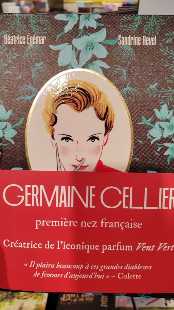 Photos de la couverture du livre  Germaine Cellier, l'audace d'une parfumeuse. La couverture représente des fleurs bleues sur un fond couleur chocolat, un dessin qui représente Germaine cellier est placé dans un macaron au centre. Le livre est entouré d'un bandeau rouge qui dit "Germaine cellier, première née française créatrice de l'iconique parfum Vent Vert"