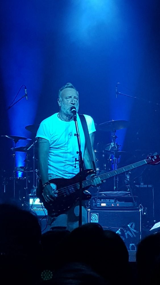 Une photo de Peter Hook en concert, prise en 2022. Il chante et il joue de la guitare basse, plongé dans une lumière bleue. Derrière lui on voit son ampli et la batterie. Au premier plan on aperçoit l'arrière de quelques têtes dans l'obscurité. 