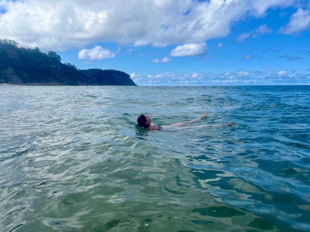 A teenage girl floating in the middle of the sea. Blue skies, fluffy white clouds, and a tree-covered cliff in the background. 