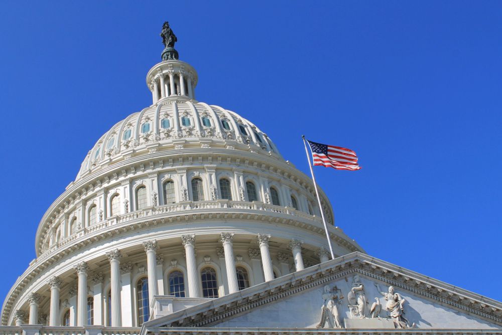 Looking up at the Capitol rotunda against a blue sky. An American flag in front of the rotunda is blowing in the wind.