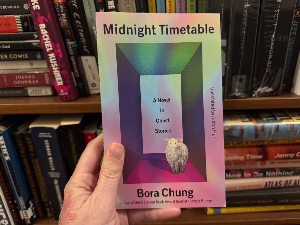 My hand, holding a copy of Bora Chung’s novel “Midnight Timetable” in front of a very full bookshelf. 
