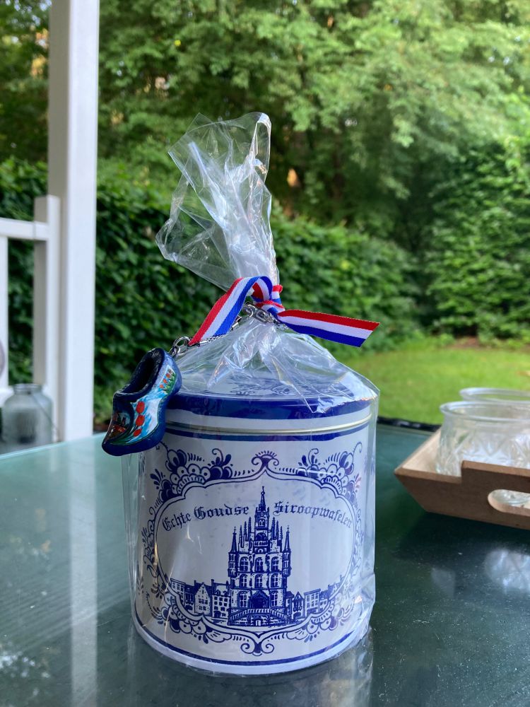 Dutch stroopwafels in a decorative tin with a clog keychain, sitting on a table with greenery in the background. 