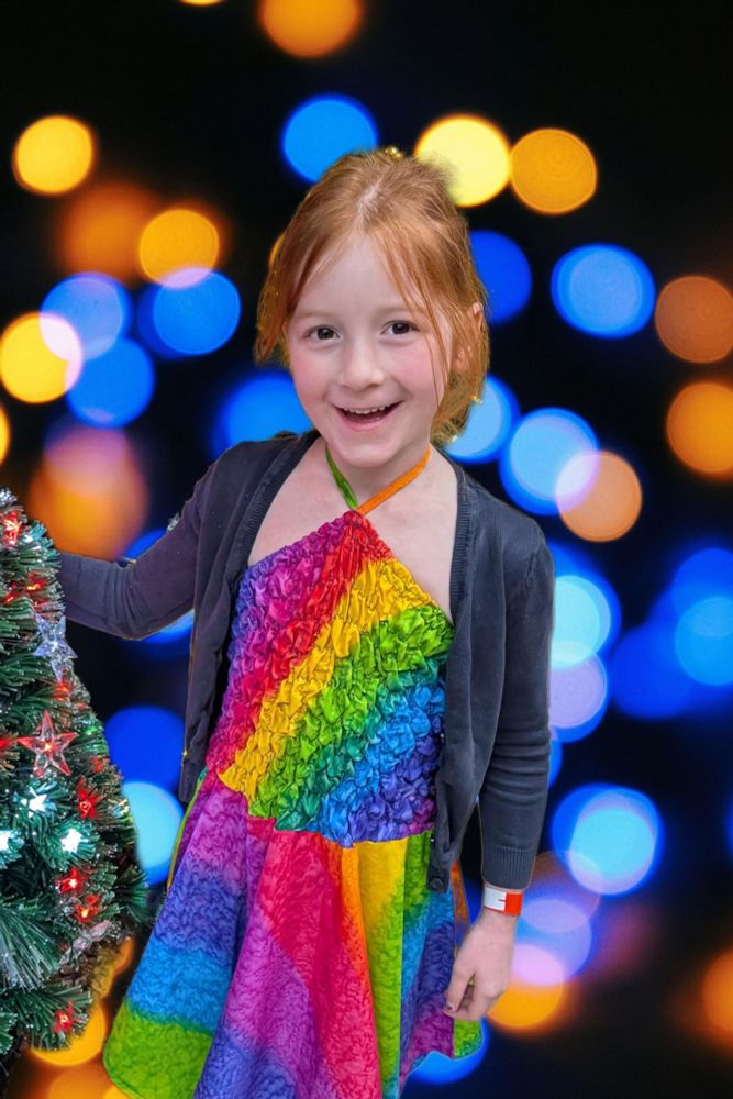 A six year old girl stands smiling wearing a rainbow dress next to a lit up Christmas tree. A stylised background of blue and orange orbs are visible.