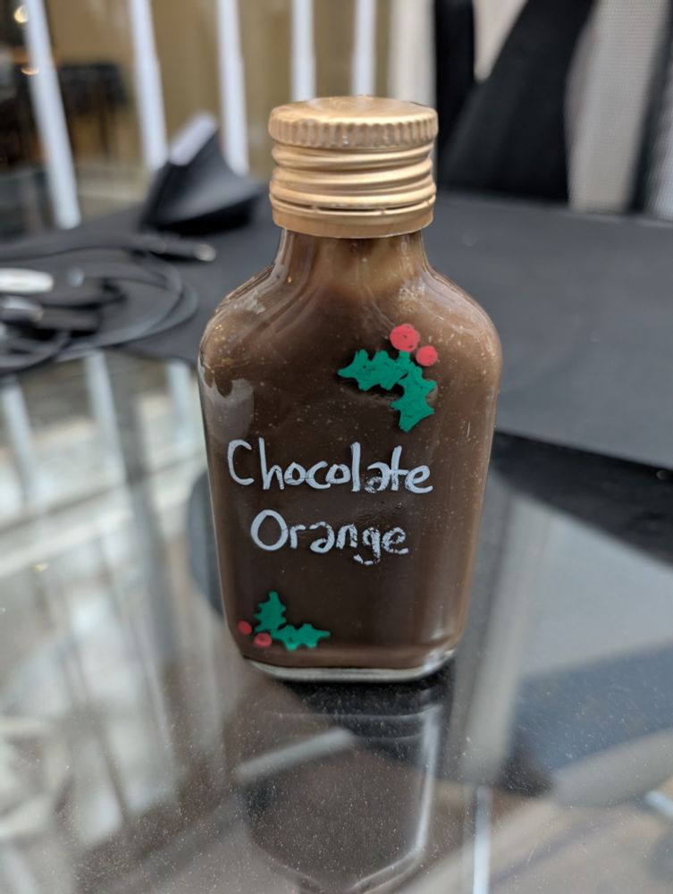 Picture of a glass bottle with 'Chocolate Orange' handwritten on the side. There are two pictures of holly and berries, also handdrawn. The bottle is filled with chocolate orange-flavoured vodka.