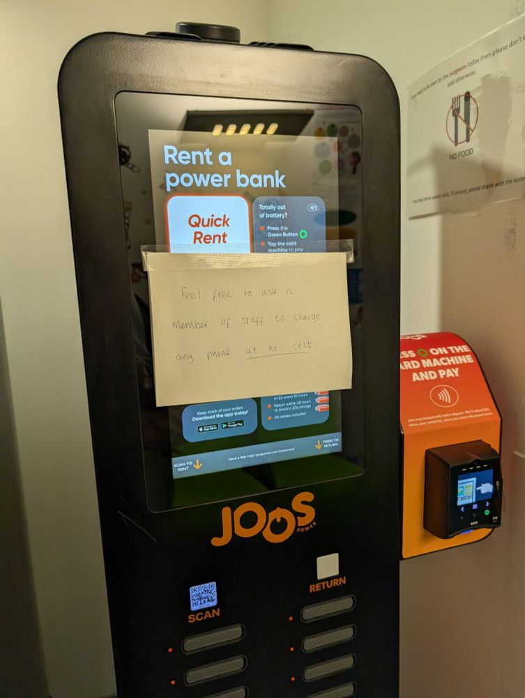 Picture of a 'rent a power bank' machine in a hospital with a handwritten note pinned to it saying: "Feel free to ask a member of staff to charge any phone at no cost"