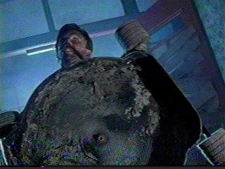 Screencap of character Lionel Cosgrove (Tim Balme) from film "Brain Dead"/"Dead Alive". He holds a push mower up, powered on, ready to slaughter some zombies.
