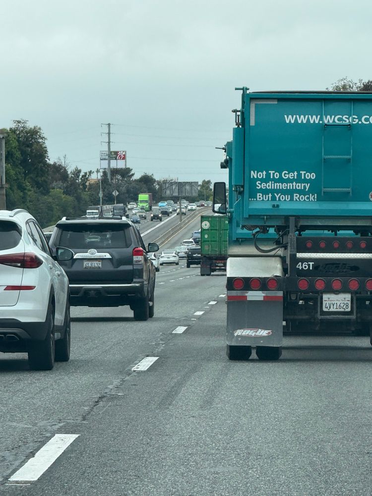 A blue semi truck on a freeway with a decal on the back that says “not to get too sedimentary but you rock!”