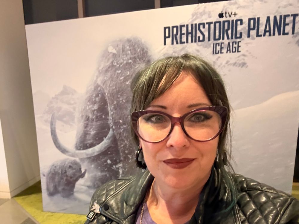 A woman with glasses and a leather jacket standing in front of a large image of a mother and baby mammoth in the snow and the words prehistoric planet ice age at the top. 