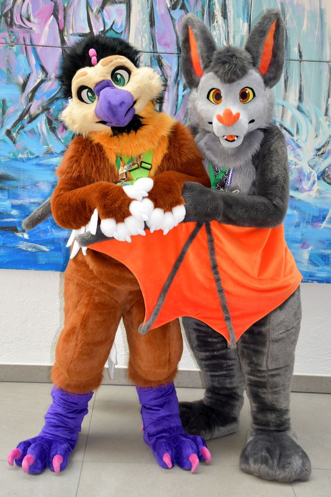An avian and bat fursuit standing together in front of a large painting