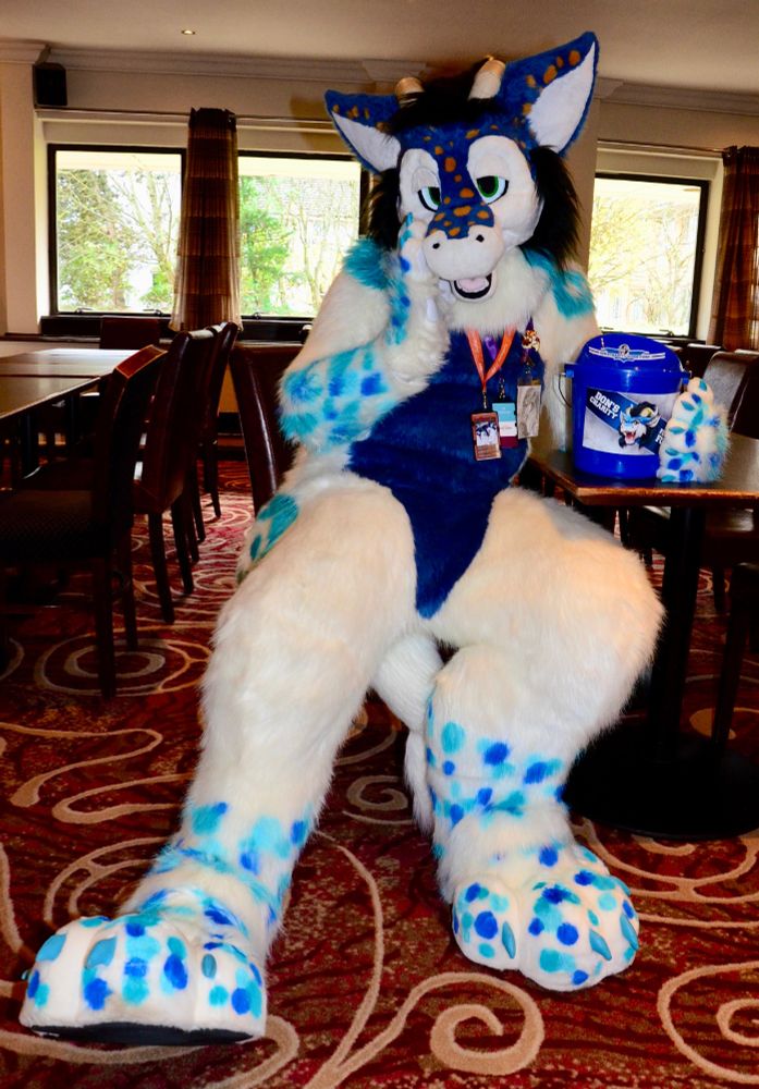 A white and blue dragon fursuit reclining in a chair in a hotel bar. 
