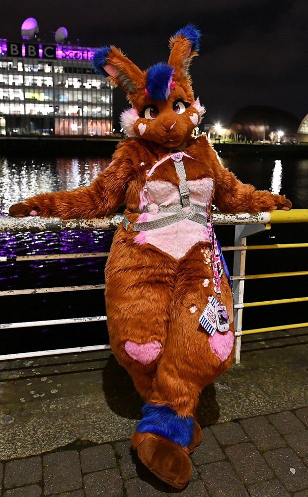 A brown and pink bunny fursuit with a heart pattern in her fur. She is wearing a harness and relaxing against a rail overlooking a river. It is night and the lights of the buildings opposite are reflected in the water. 