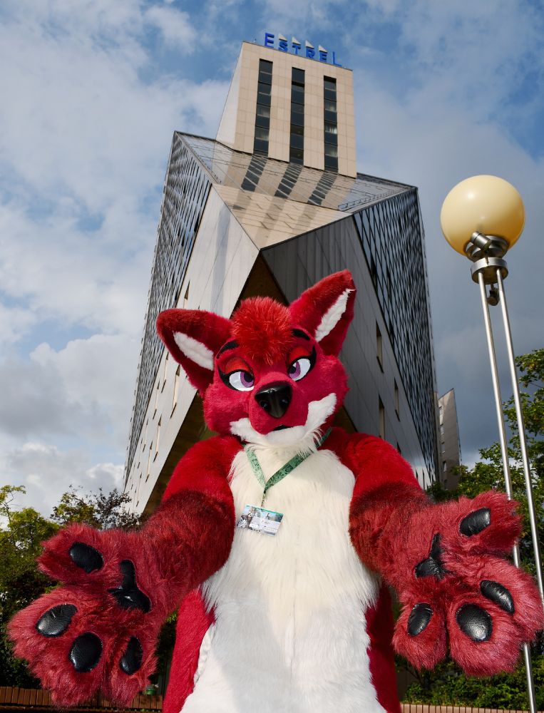 A red vixen fursuit extending her paws towards the camera with a high rise hotel in the background.