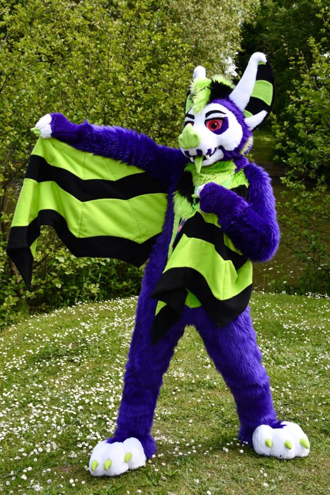 A purple & green bat fursuit posing with a wing extended by a lakeside. There is green foliage all around. 