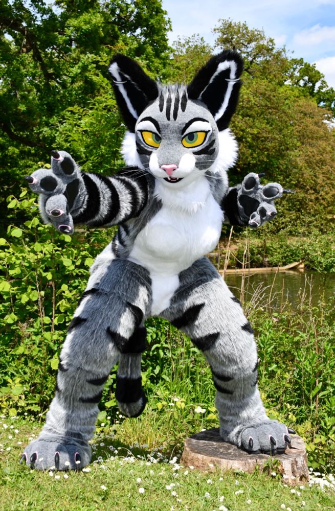 A tabby cat fursuit with bright yellow eyes posing by a lakeside with a clawed paw extending towards the camera. There is green foliage all around. 