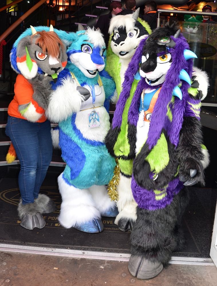 Four fursuiters gathering for a group photo outside a bar in Manchester. A grey demon goat, a blue & white bunny/goat, a green goat, and a purple & green monster.