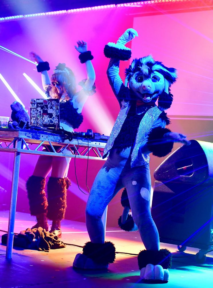 A poodle fursuit wearing a sparkly waistcoat on stage with dance lighting all around. The DJ, dressed as a poodle, is in the background with arms raised.