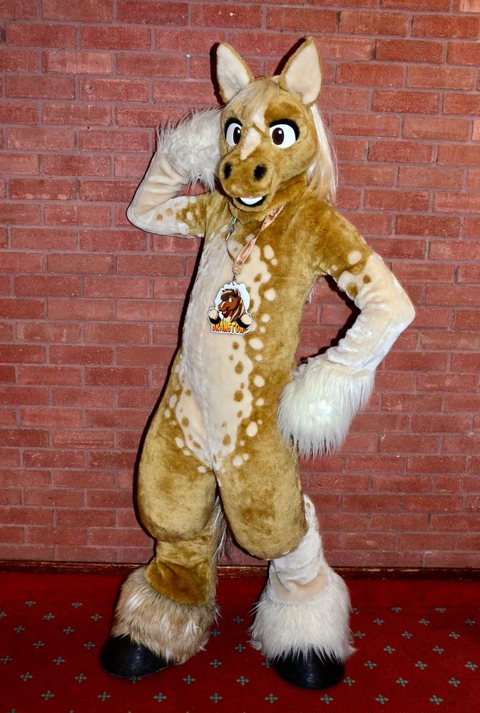 A sandy coloured horse fursuit posing in front of a brick wall.