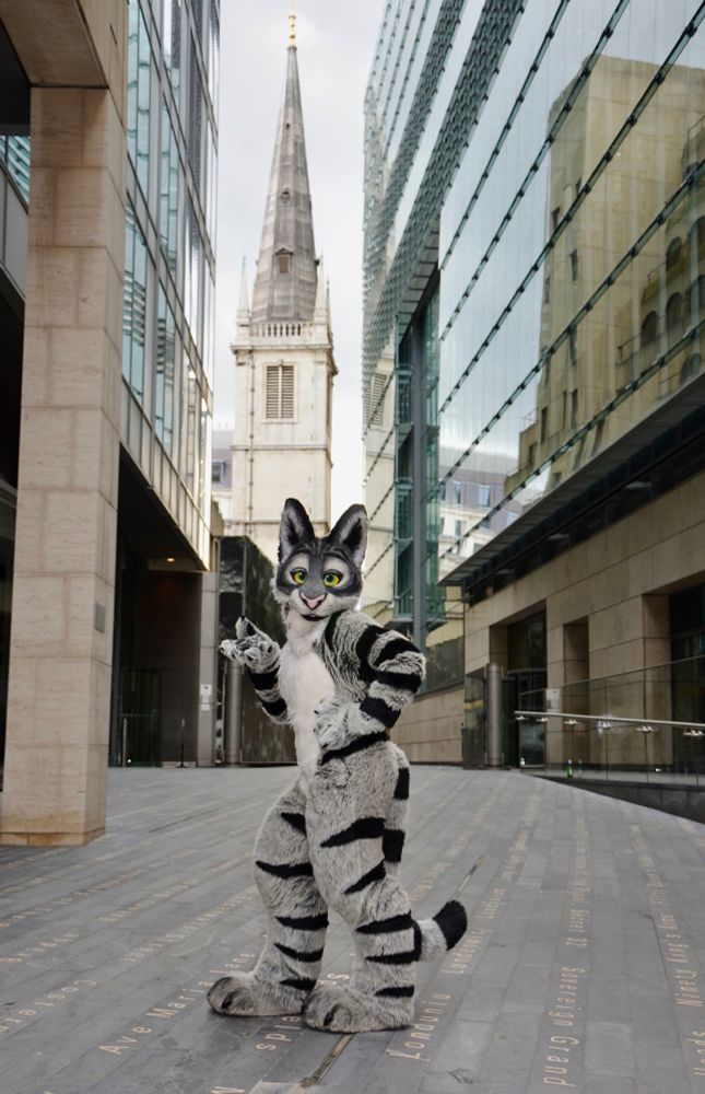 A tabby cat fursuit posing in an alleyway in London.
There is a church in the background and glass fronted buildings to each side.