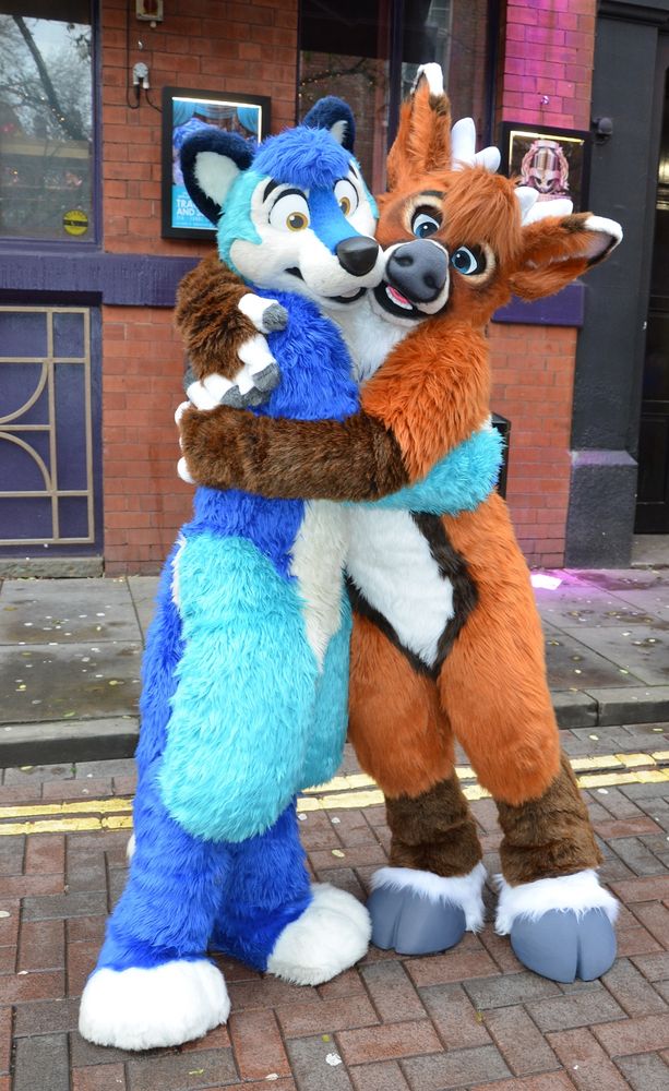 A blue wolf fursuiter posing with a brown deer fursuiter. They are on a street with a pub in the background.
