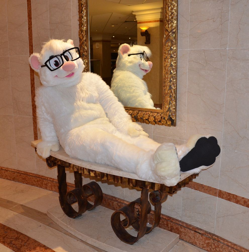 A white meerkat fursuit wearing oversized glasses and sitting on a bench in front of a mirror