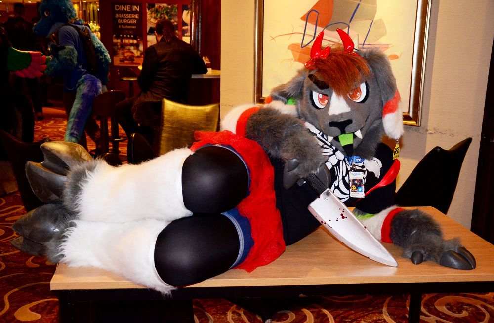 A demon goat fursuit wearing devil horns, reclining on a table and holding a large prop knife