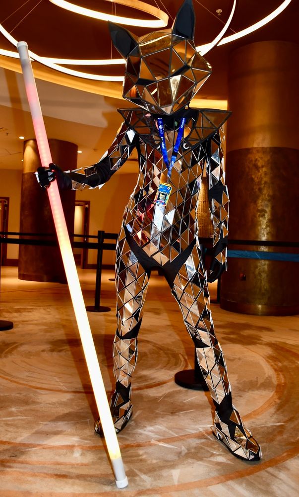 A shiny mirrored fox costume holding an illuminated staff. He is posing in a hotel lobby.