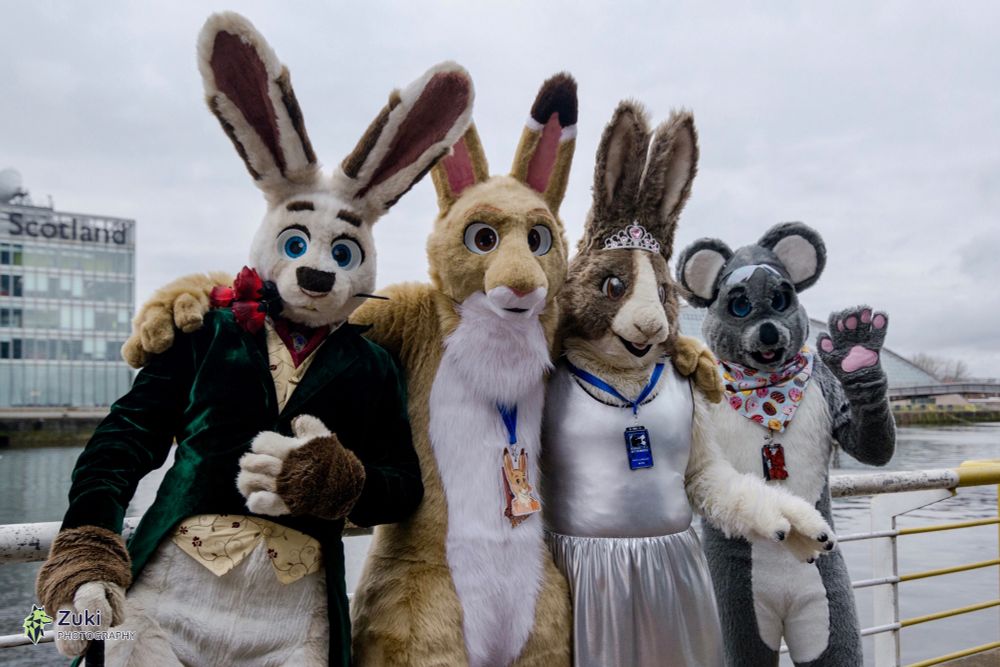 A photo of four fursuiters standing by a rail at the banks of the River Clyde in Glasgow. There is a white rabbit wearing a fancy waistcoat and with a rose in his mouth, there is a brown and white hare, a brown and white rabbit wearing a silver dress and tiara, and a grey koala. 