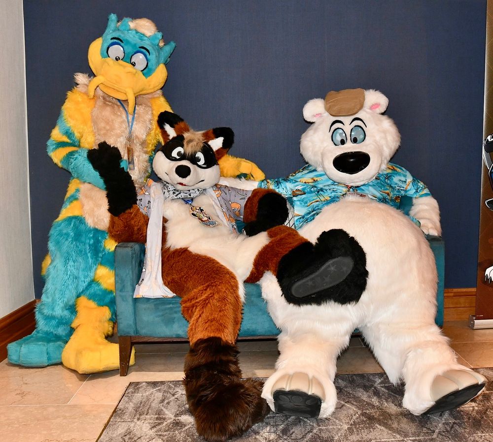 Three fursuiters - a dragon, a fox, & a polar bear, posing around a sofa.