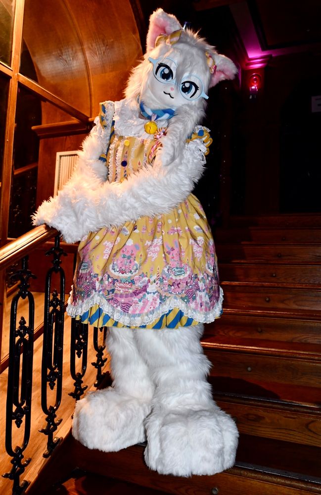 A white feline fursuit wearing a bright yellow patterned dress, standing on a wooden staircase.