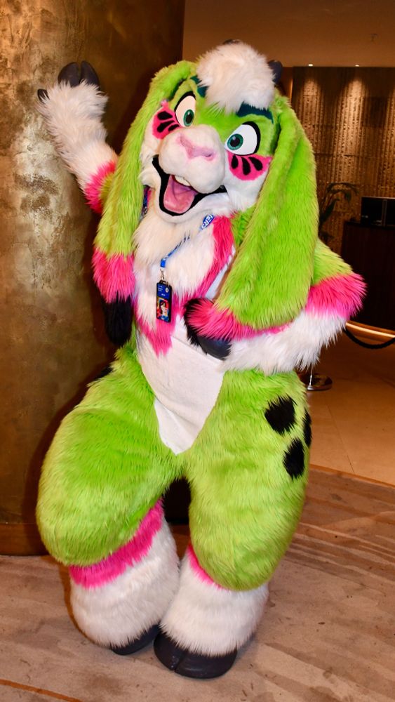 A green, white, & pink goat fursuit posing by a gold pillar in a hotel lobby.