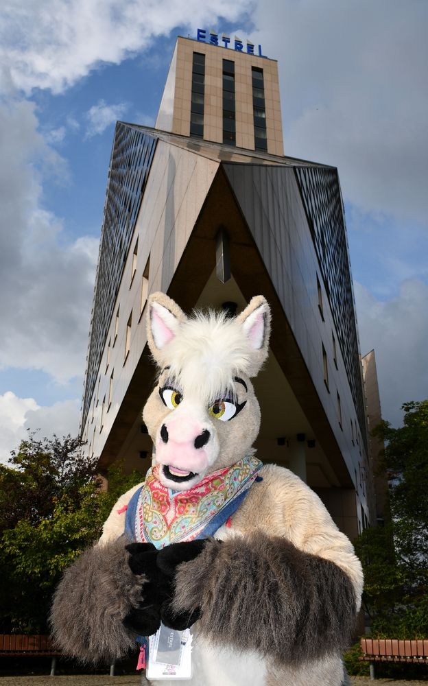 A sandy coloured horse fursuit with a high rise hotel in the background.