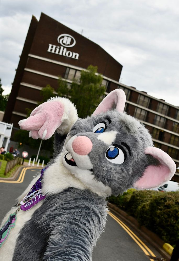 A closeup of a rat fursuit posing with a hotel in the background.