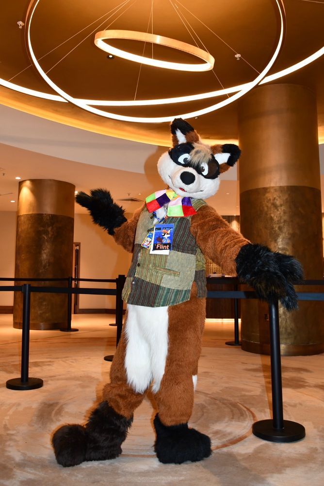 A fox fursuit wearing a patched waistcoat and bow tie posing in a hotel lobby.