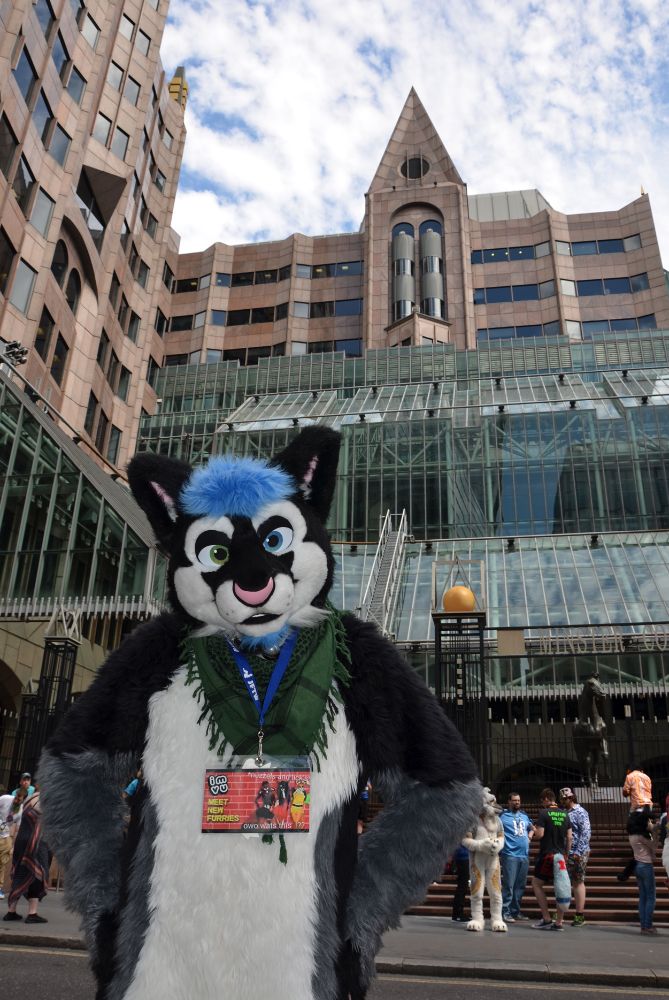 A white & black fursuiter posing with a glass fronted building in the background.