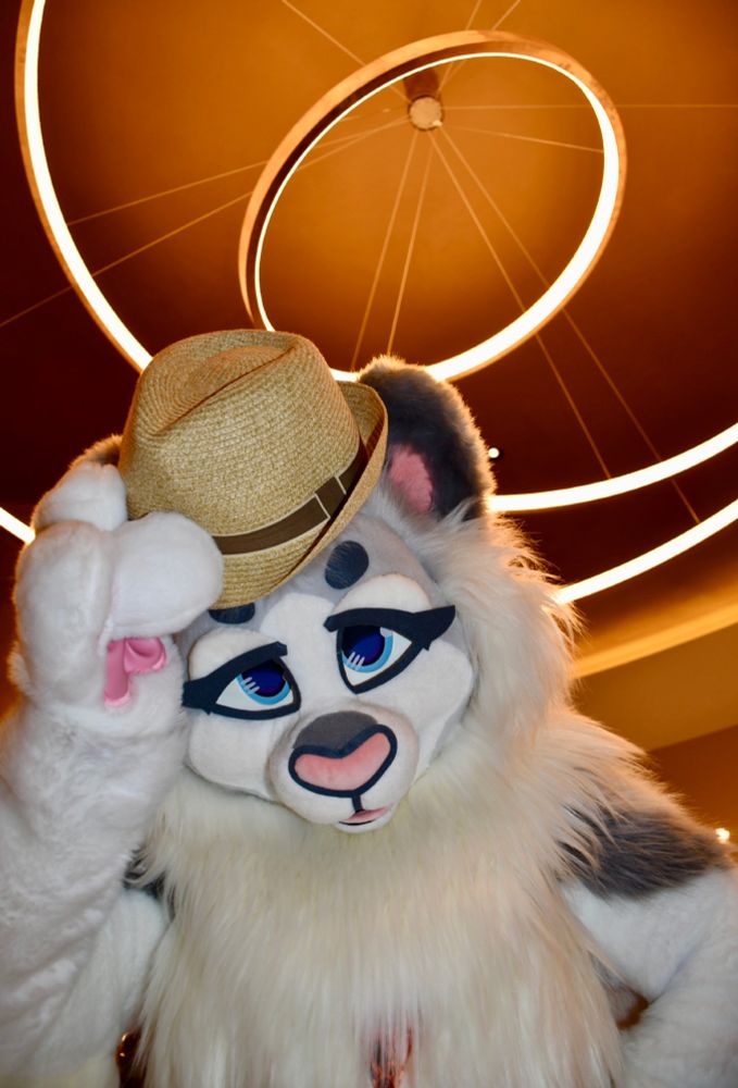 A closeup of a white feline fursuit with a hat in their paw. They are standing under some hotel lobby lighting.