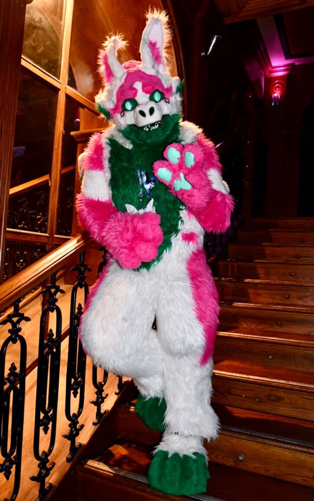A multicoloured ( red, white, green ) monster fursuit standing on some wooden stairs.