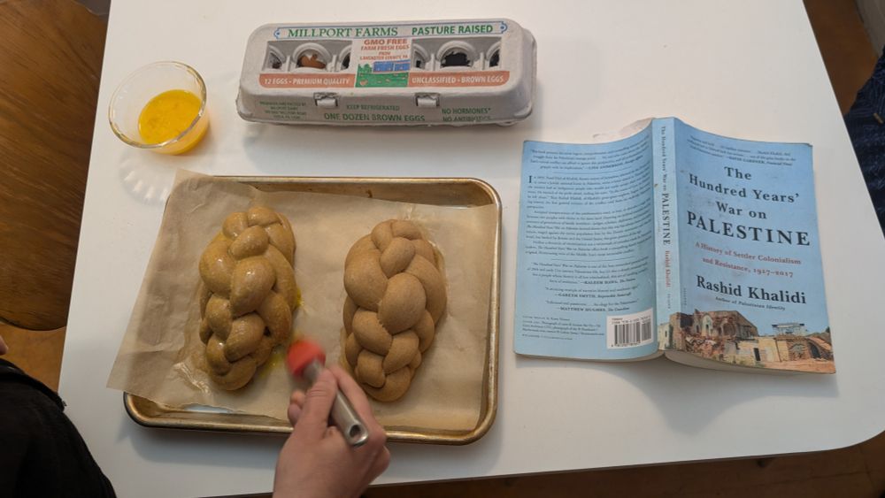 two loaves of challah being basted, and next to them on the table is the book The 100 Years' War on Palestine by Rashid Khalidi, open and spine up.