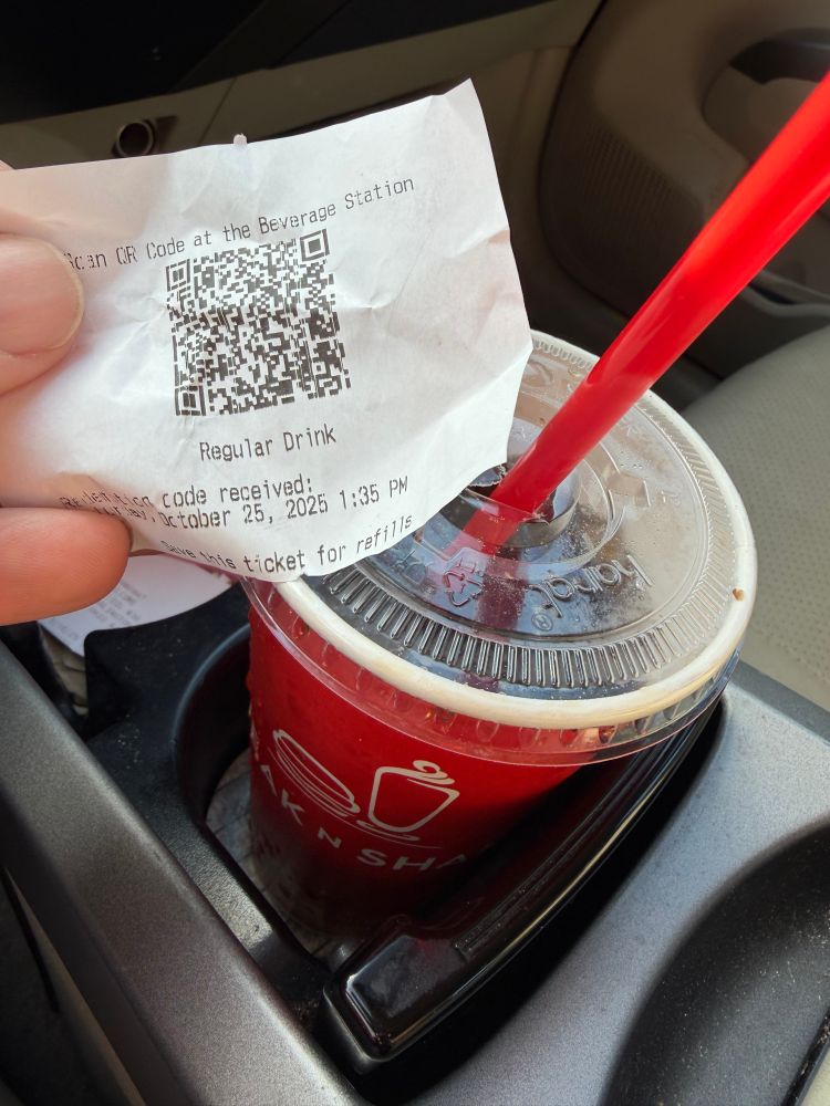 A soft drink cup sits in a car cup holder. A hand holds a receipt near it with a QR code on it.