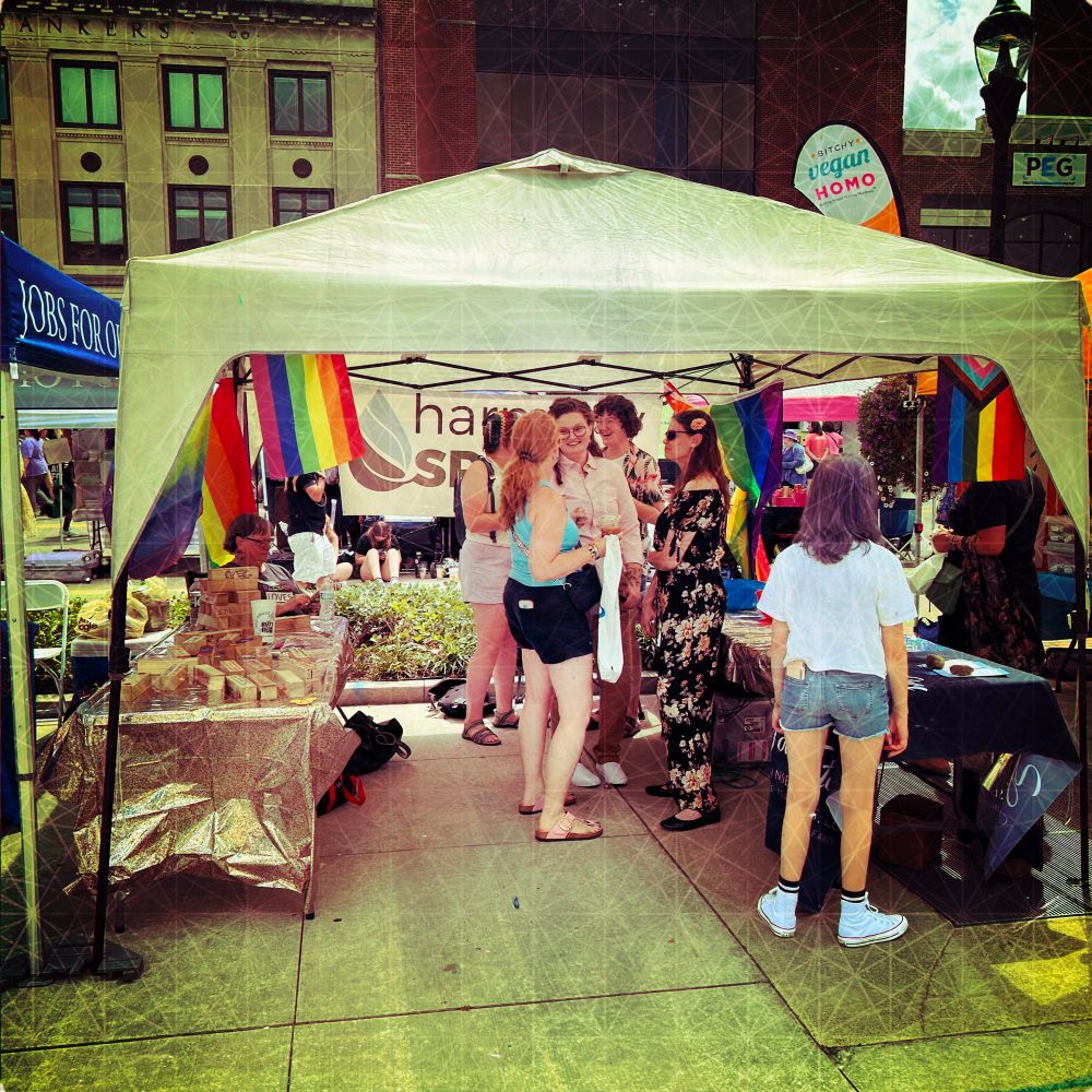 People stand under a pop-up tent on a city street. There are lots of rainbow flags around d. 