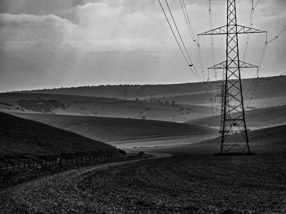 Black and white image, winding valley through the Downs, shafts of sunlight, electricity pylon. 