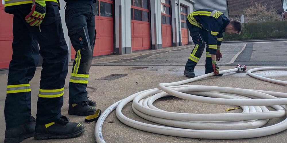 Ein Feuerwehrschlauch liegt in einem Loop auf dem Boden. Daneben stehen zwei Feuerwehrleute, deren Beine man sieht. Ein dritter Feuerwehrmann beugt sich nach unten, um das Strahlrohr anzuheben.