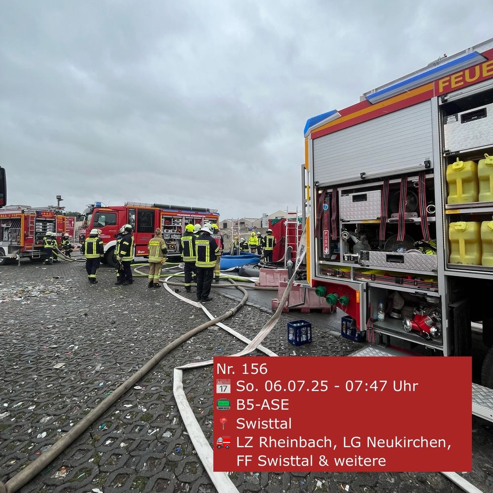 Seitenansicht eines Feuerwehrautos. Die Rollladen der Geräteräume sind geöffnet. Im Hintergrund stehen Feuerwehrleute.