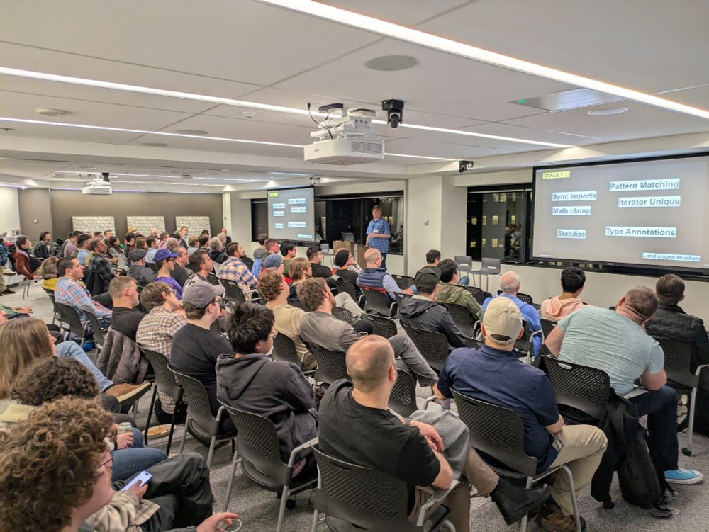 A room full of people facing a speaker at a podium presenting slides on a screen about JavaScript features 