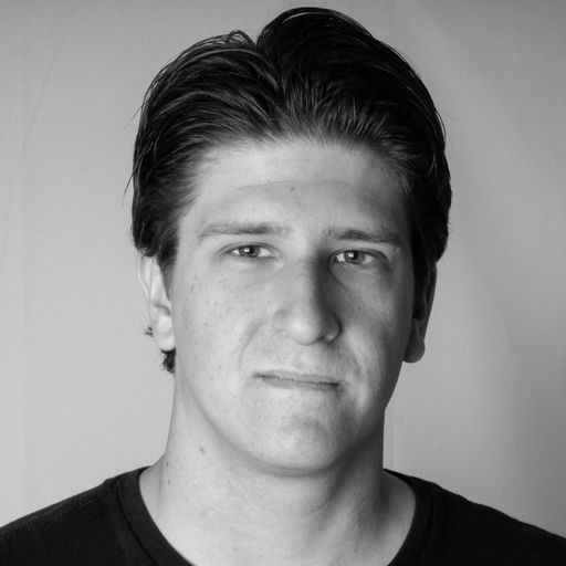 Black and white portrait of Kenny with a serious expression in a black t-shirt. The background is matte gray and Kenny's hair is styled up.