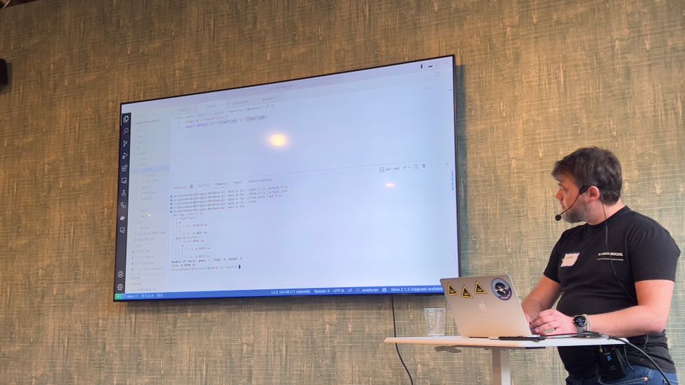 Sergey is standing by the podium showing VSCode with his functionalscript tests running.