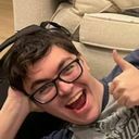 Will smiling excitedly with his mouth open and a big thumbs up. Will is laying sideways and has a couch behind him.