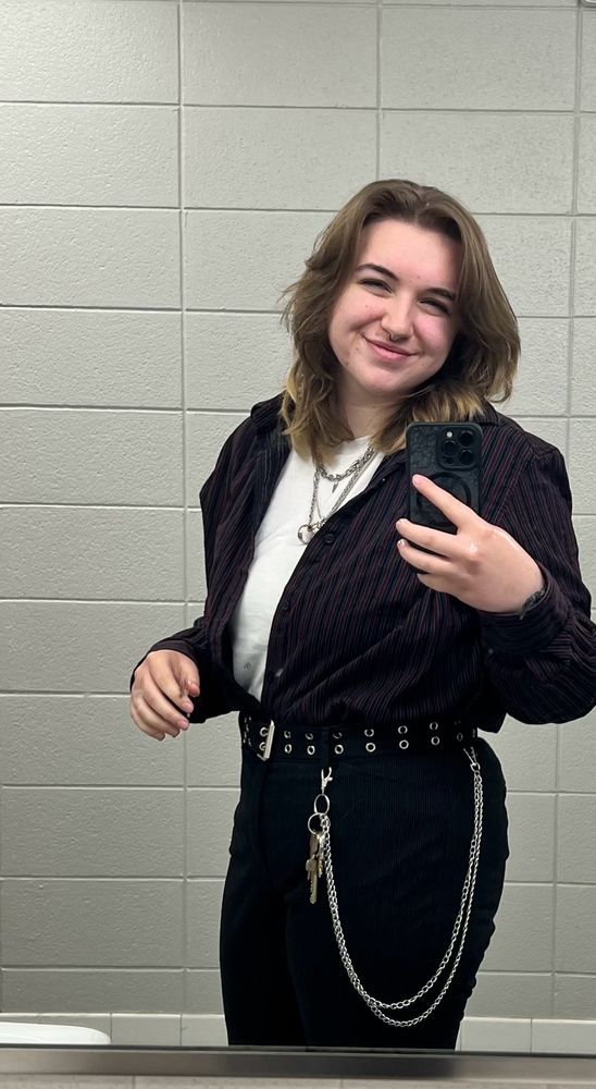 Me in front of a mirror wearing a white undershirt with a dark striped button-up and black pants. there are silver necklaces and a silver wallet chain.
