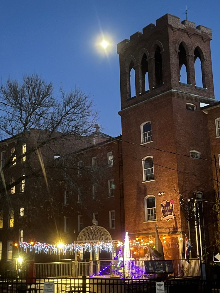 Old mill building at dusk with castle-like tower on the right, strings of holiday lights at the bottom, leafless trees on the left, and a bright moon above. 