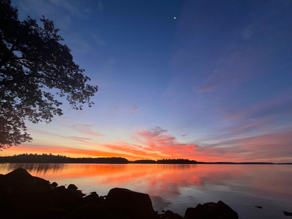 Spectacular sunrise over a lake, with a branch visible on the left, black rocks along the bottom, deep orange clouds along the horizon, and dark blue sky at the top. 